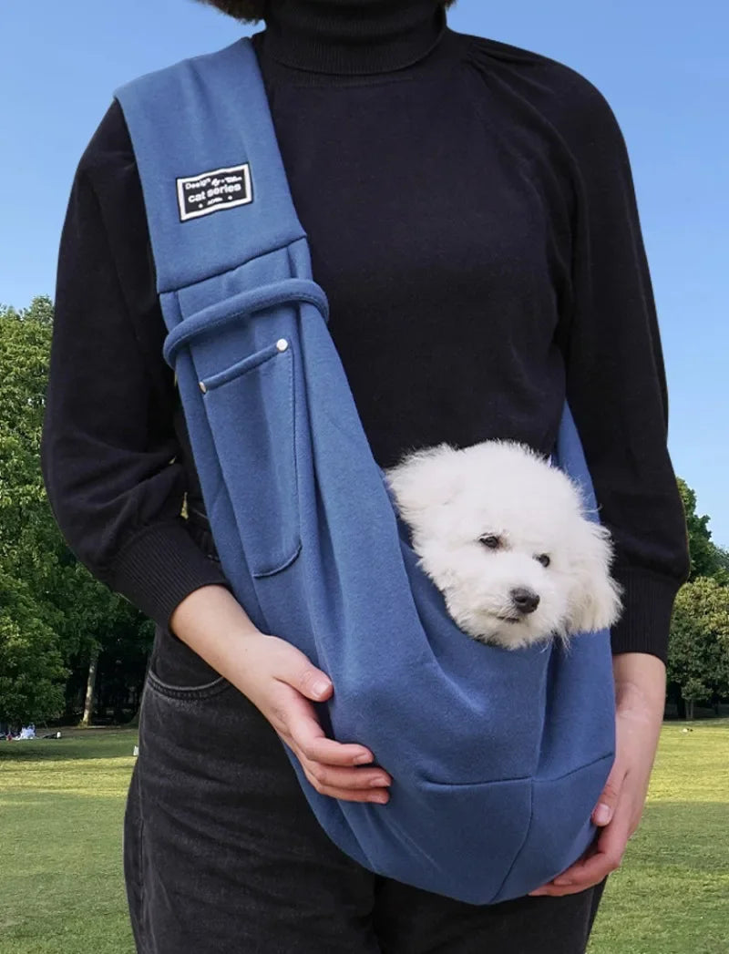 Person holding a blue pet sling with a small white dog inside, outdoors on a sunny day.