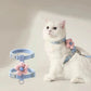White cat wearing a light blue harness with pink floral attachments on a plain background