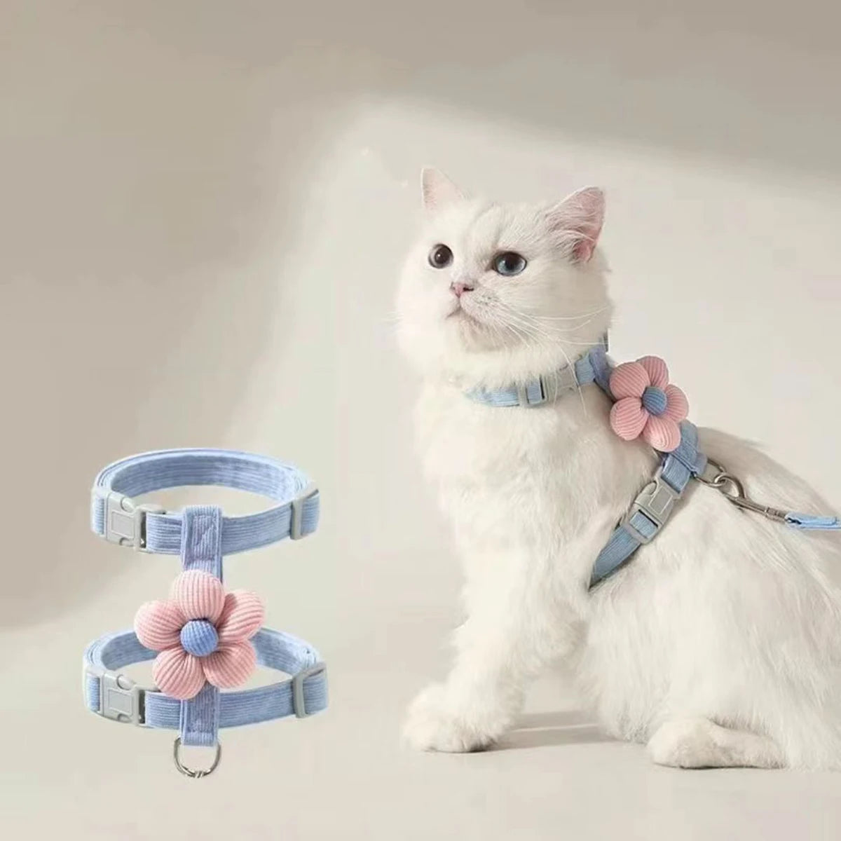 White cat wearing a light blue harness with pink floral attachments on a plain background