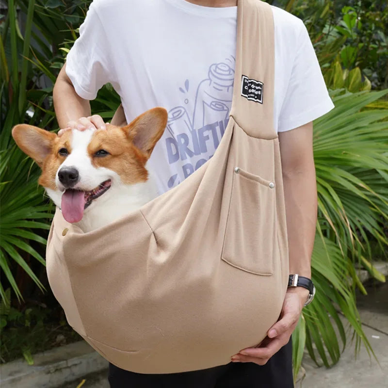 Person holding a corgi in a beige sling with greenery in the background