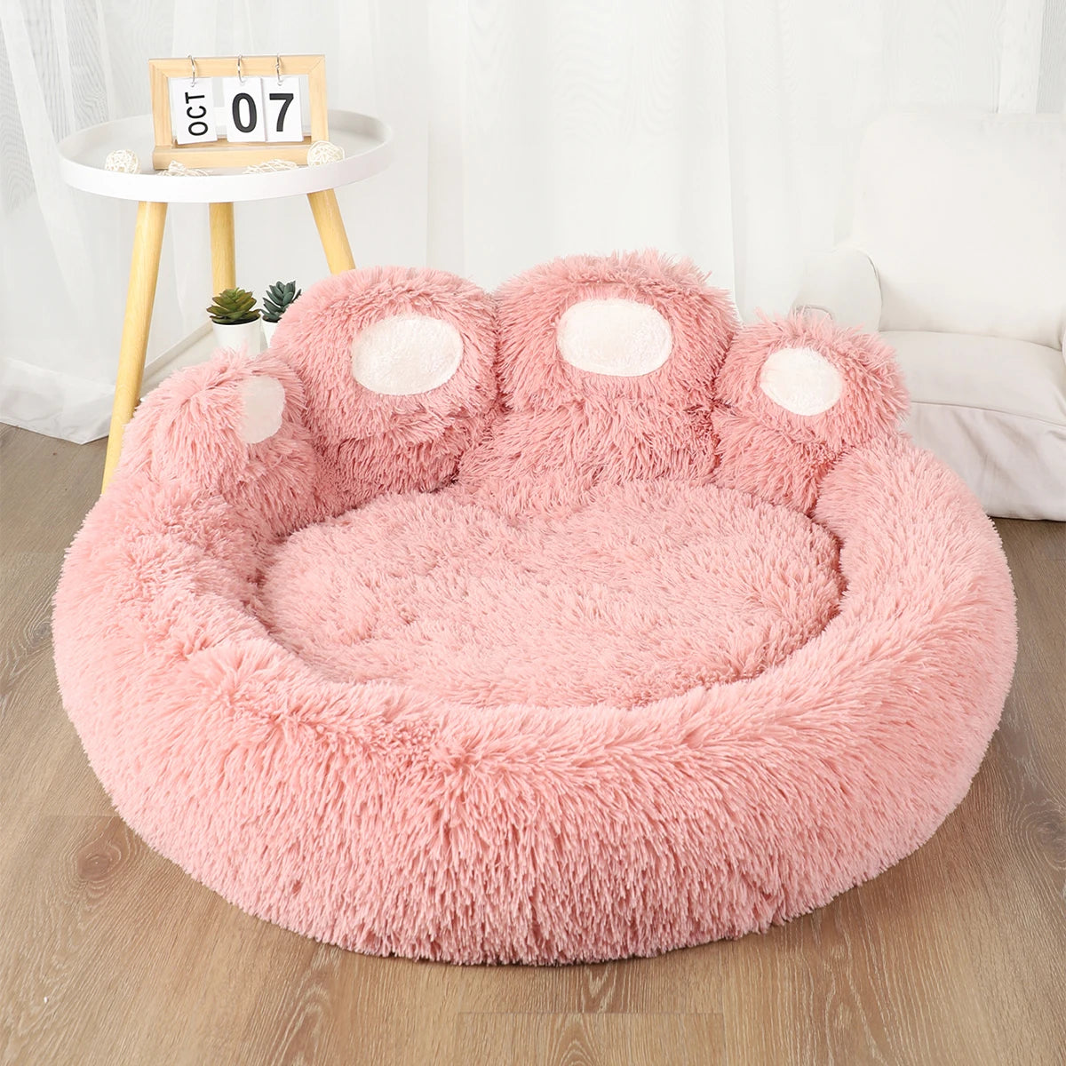 Pink paw-shaped pet bed on a wooden floor with a white curtain in the background.