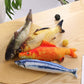 Four fish-shaped toys on a wooden surface with a straw hat and small items.