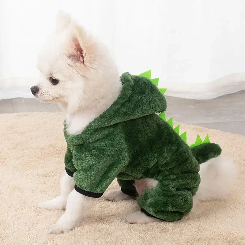 Small white dog wearing a green dinosaur costume on a beige surface.