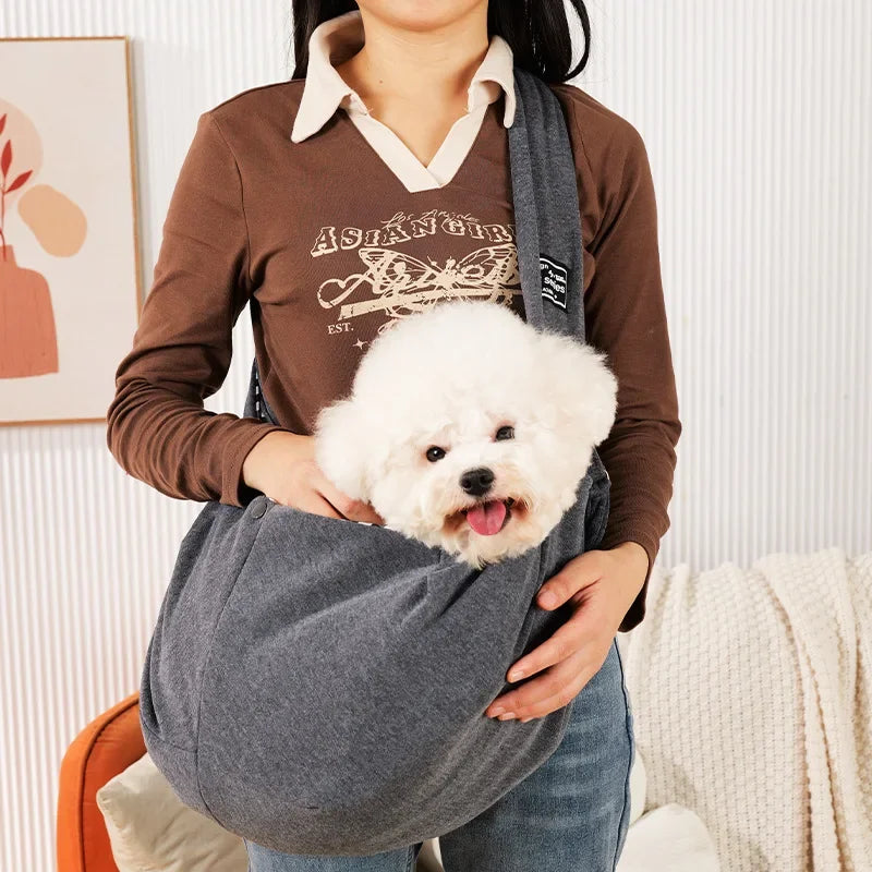 Person holding a small white dog in a gray pet sling indoors.