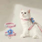 White cat wearing a pink harness with a blue flower attachment on a light background