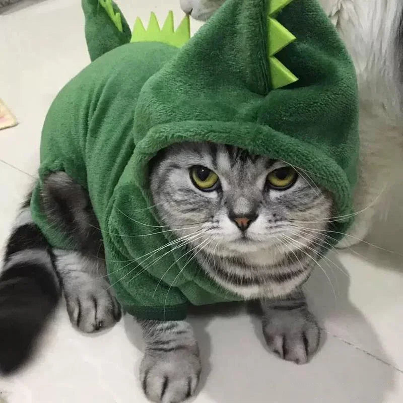 Cat wearing a green dinosaur costume on a light-colored floor.