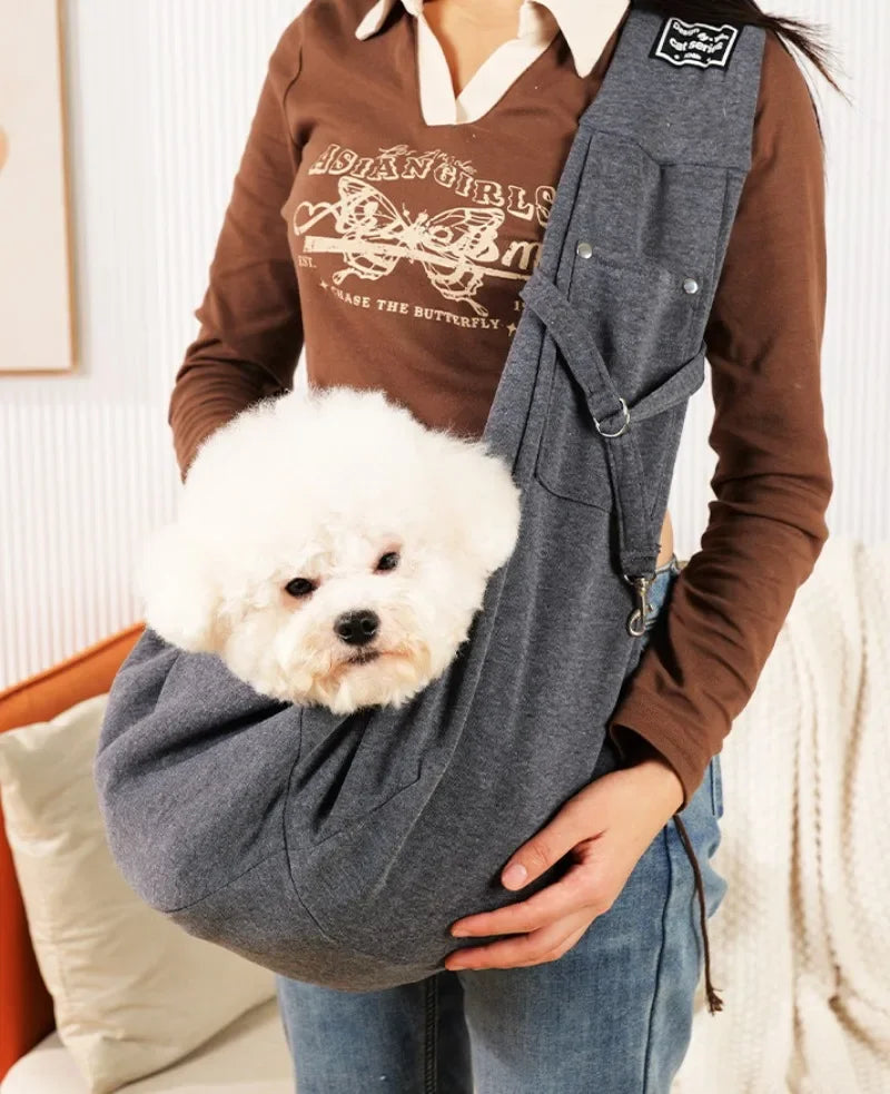 Person holding a small white dog in a gray sling, wearing a brown shirt with graphic design.