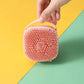Pink silicone scrubber held by a hand on a yellow and green background
