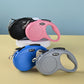 Four retractable dog leashes in black, pink, blue, and gray on a beige surface with a light blue background.